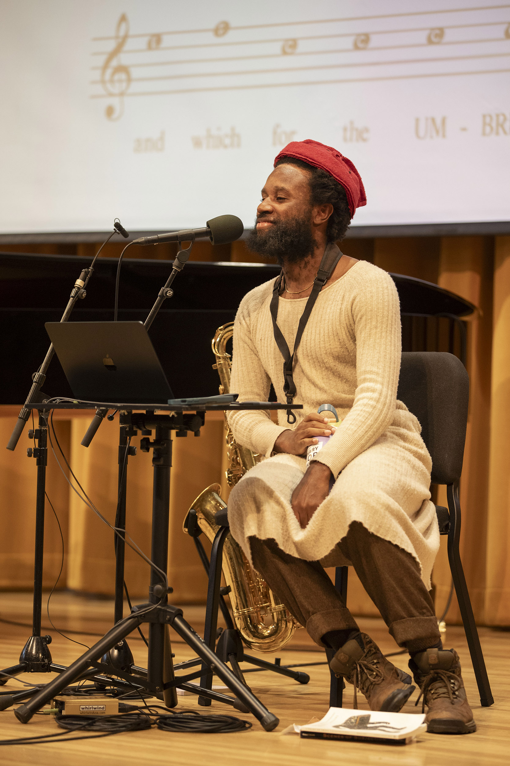 Jerome Ellis sits and speaks into microphone on Paine Hall stage with piano, saxophone, and laptop. A screen behind him projects poetry and sheet music.