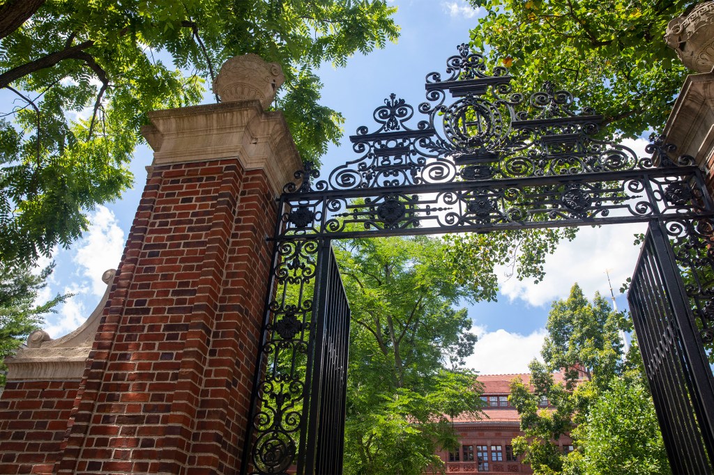 Sever Hall and a gate with an “H” is framed by foliage in Harvard Yard.