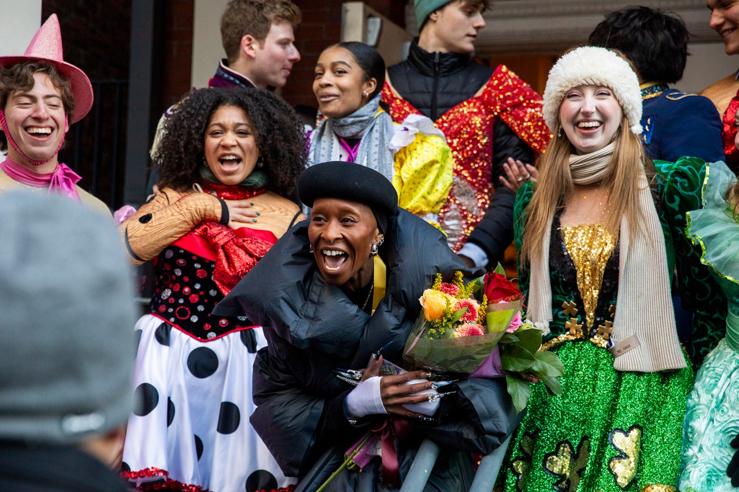 Cynthia Erivo, holding a bouquet of flowers, is surrounded by festively costumed cast members of Hasty Pudding Theatricals.