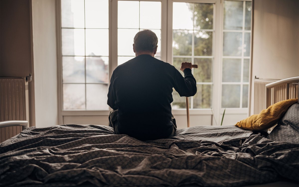 A lonely elderly man is sitting on the bedlooking out the window.