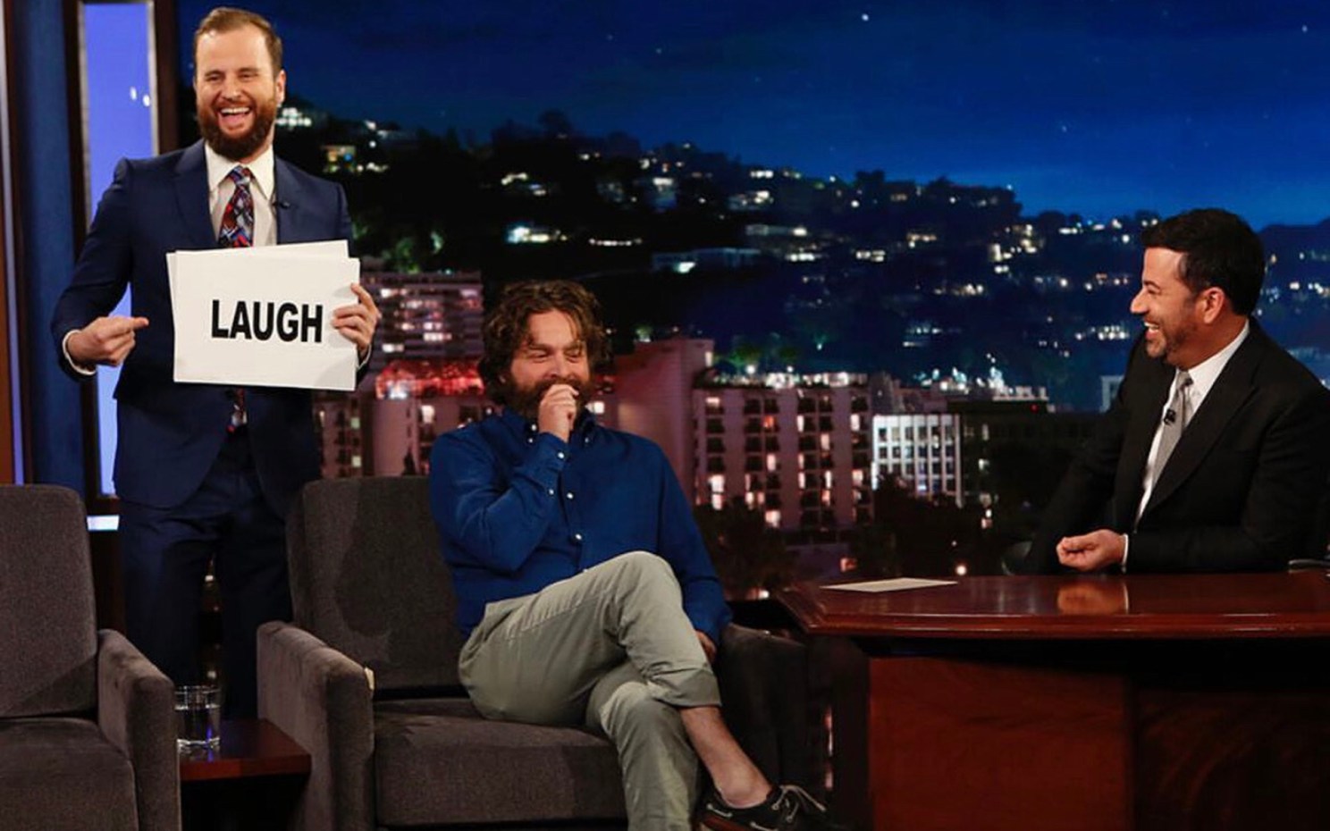 Will Burke holds a "laugh" sign as Zach Galifianakis is interviewed by Jimmy Kimmel.