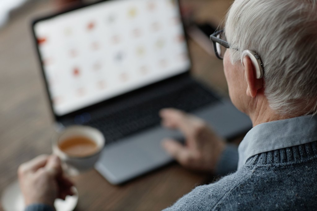 Over shoulder shot of older man wearing hearing aid while working on laptop.