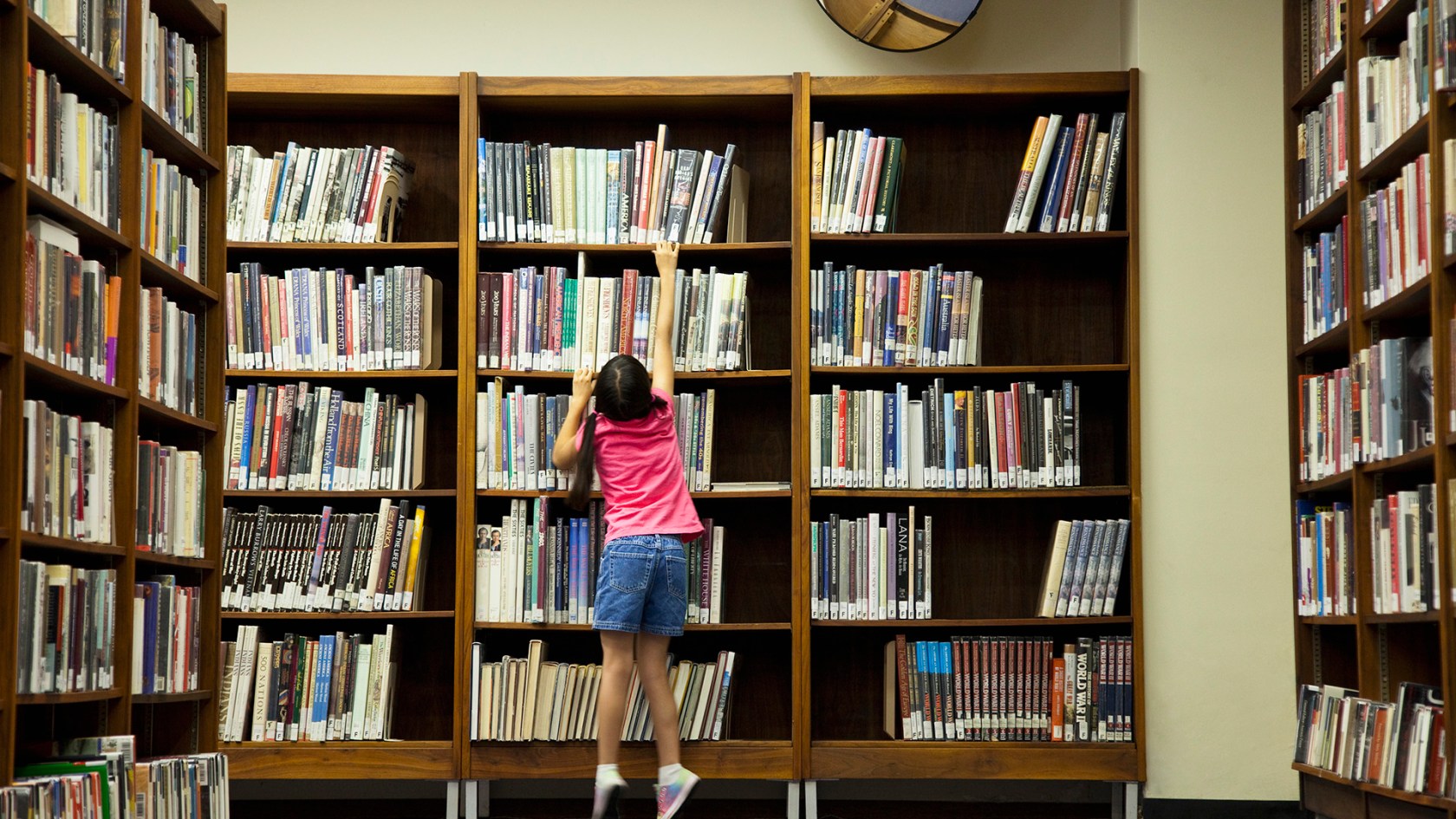 Child stands on tip-toes to reach for a book off a shelf.