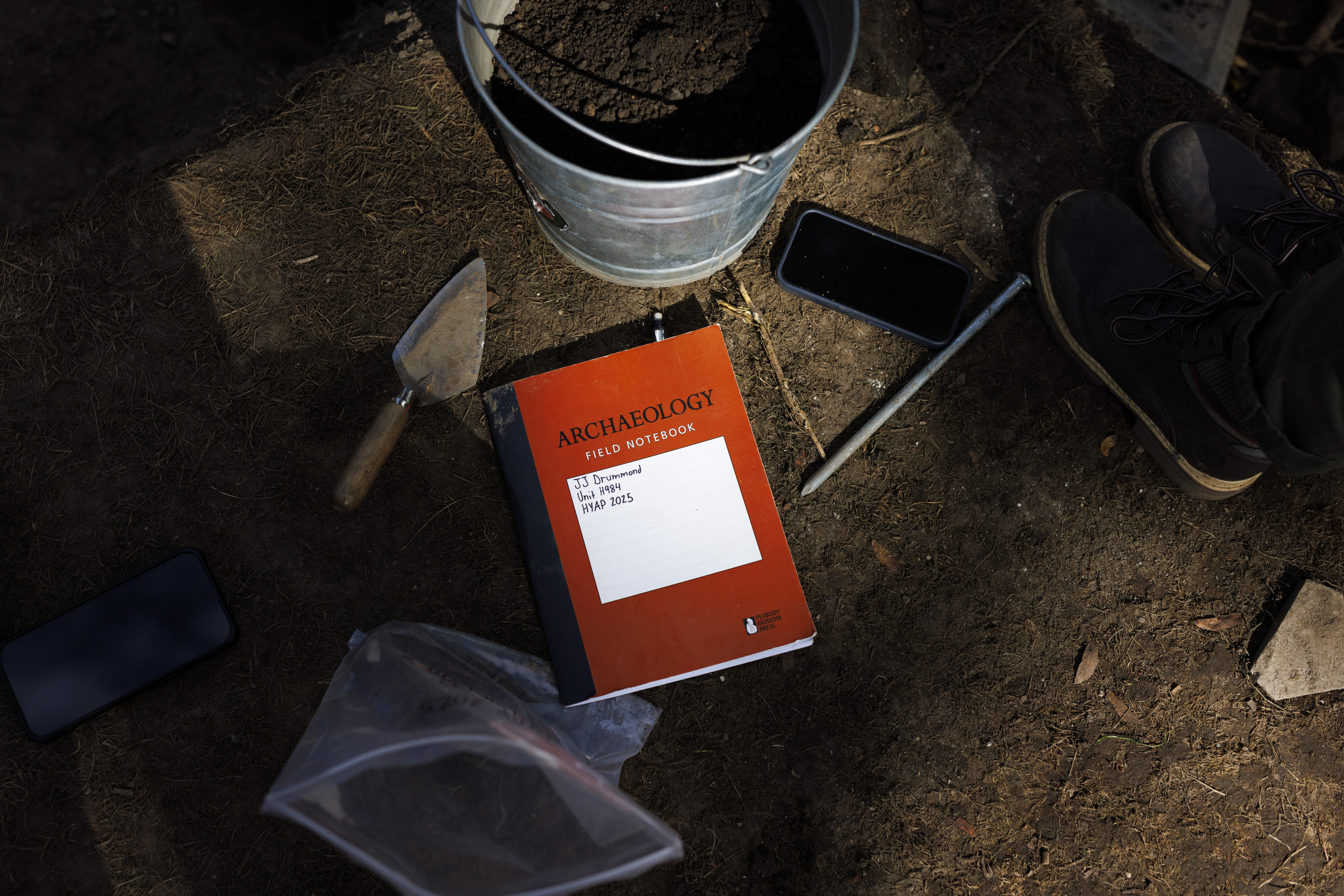 Tools of the trade, including a field notebook, bucket, trowel, and artifact bag sit at the edge of the dig site.