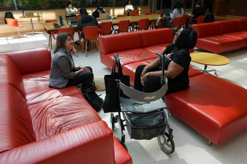 Two women sitting facing one another on couches in Smith Campus Center. One woman is smiling and the other is leaning back laughing. A walker mobility device is in the foreground.